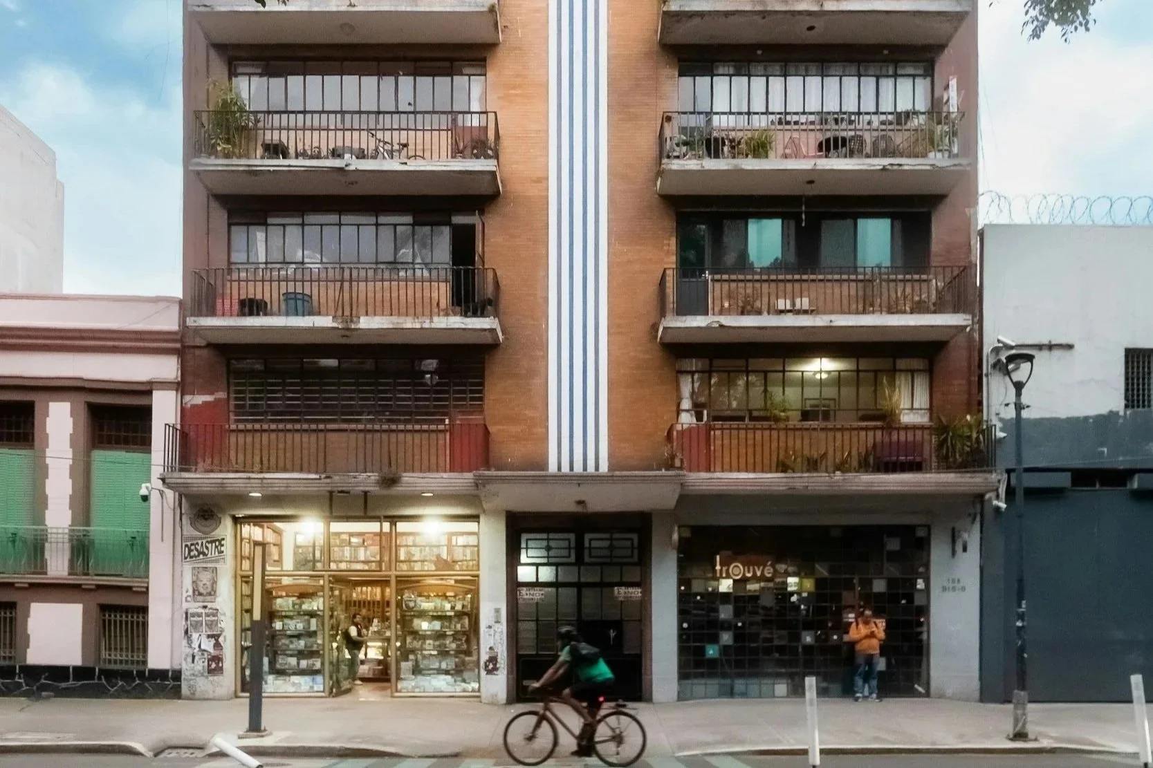 Four-story brick building with retail stores on the ground floor and apartments on the upper floors, surrounded by street lamps and a cyclist passing by.