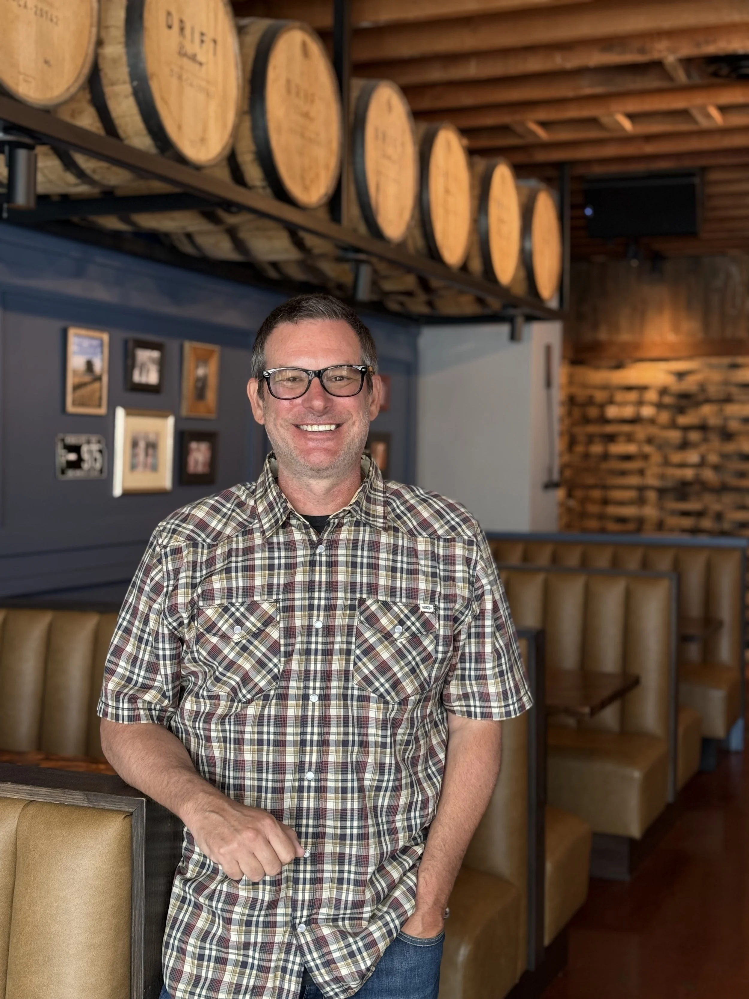 A smiling man wearing glasses and a plaid shirt standing in a restaurant with wooden barrels above and booths with leather seats.