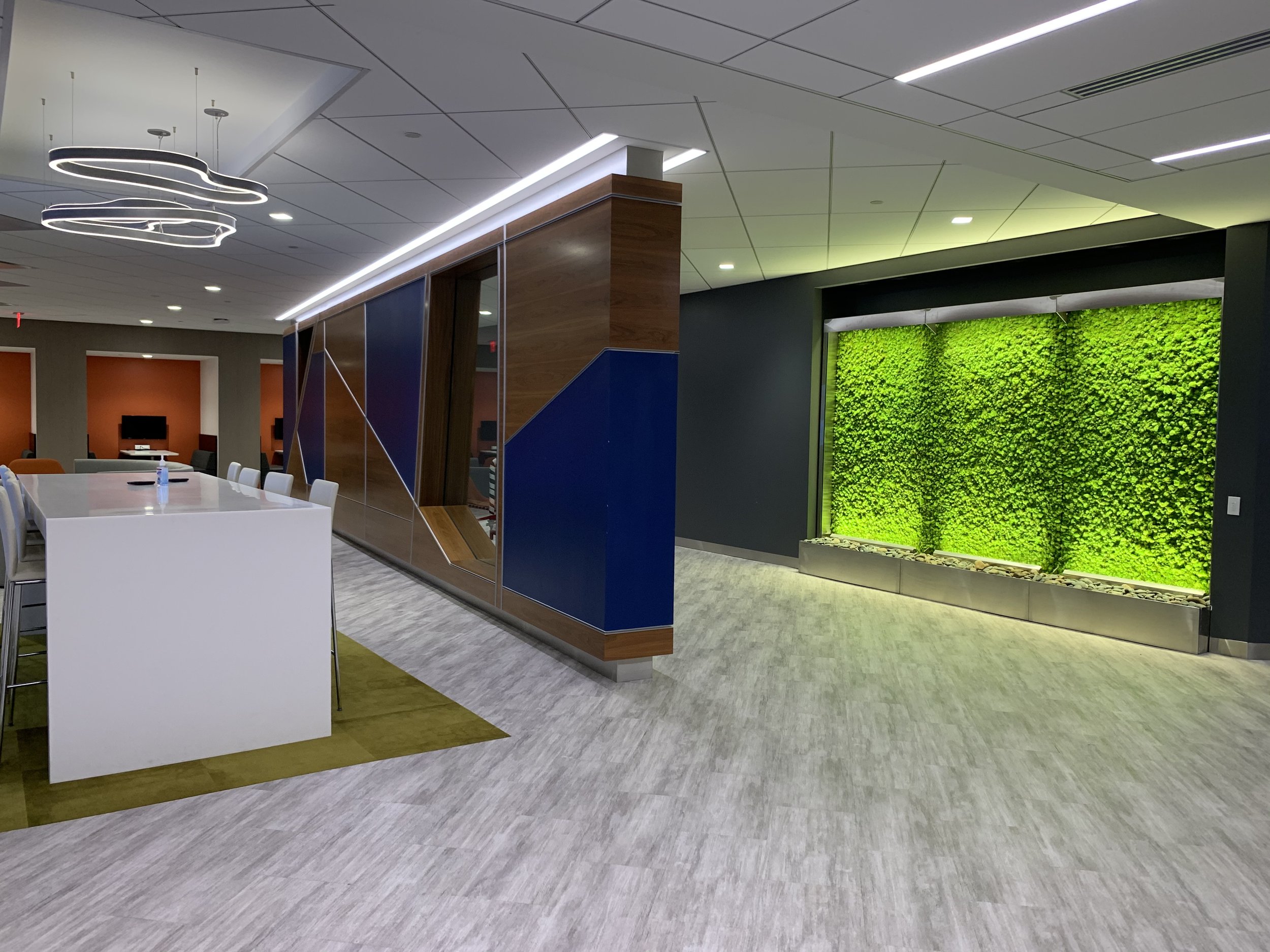 Modern office or lobby area featuring a white reception or high table, seating, a decorative ceiling light, a green living wall, and contemporary design elements.