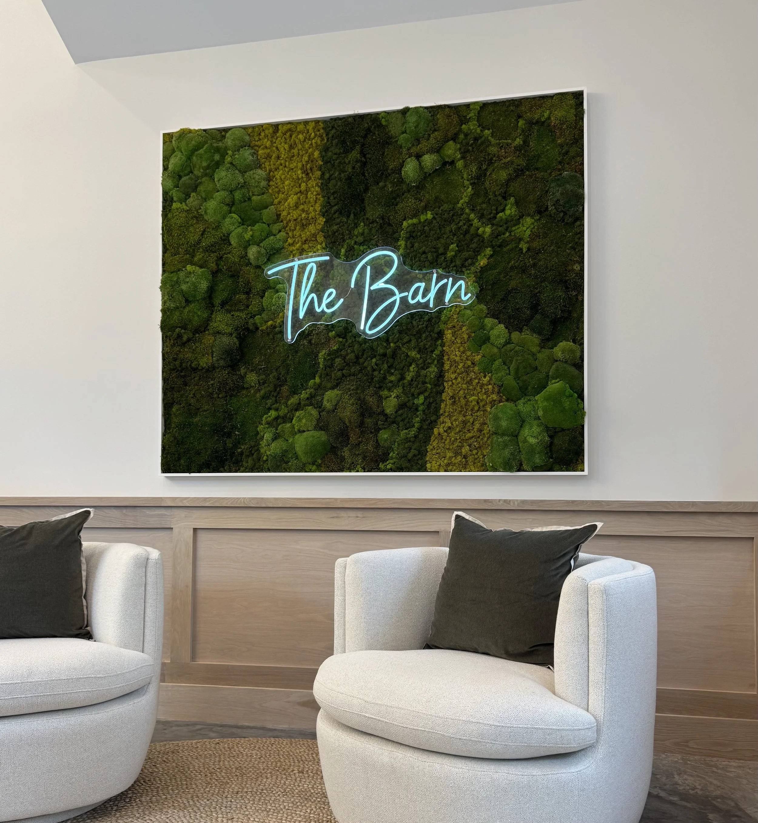 Interior of a modern room with two white armchairs, black pillows, and a green moss wall art piece with neon sign reading 'The Barn'