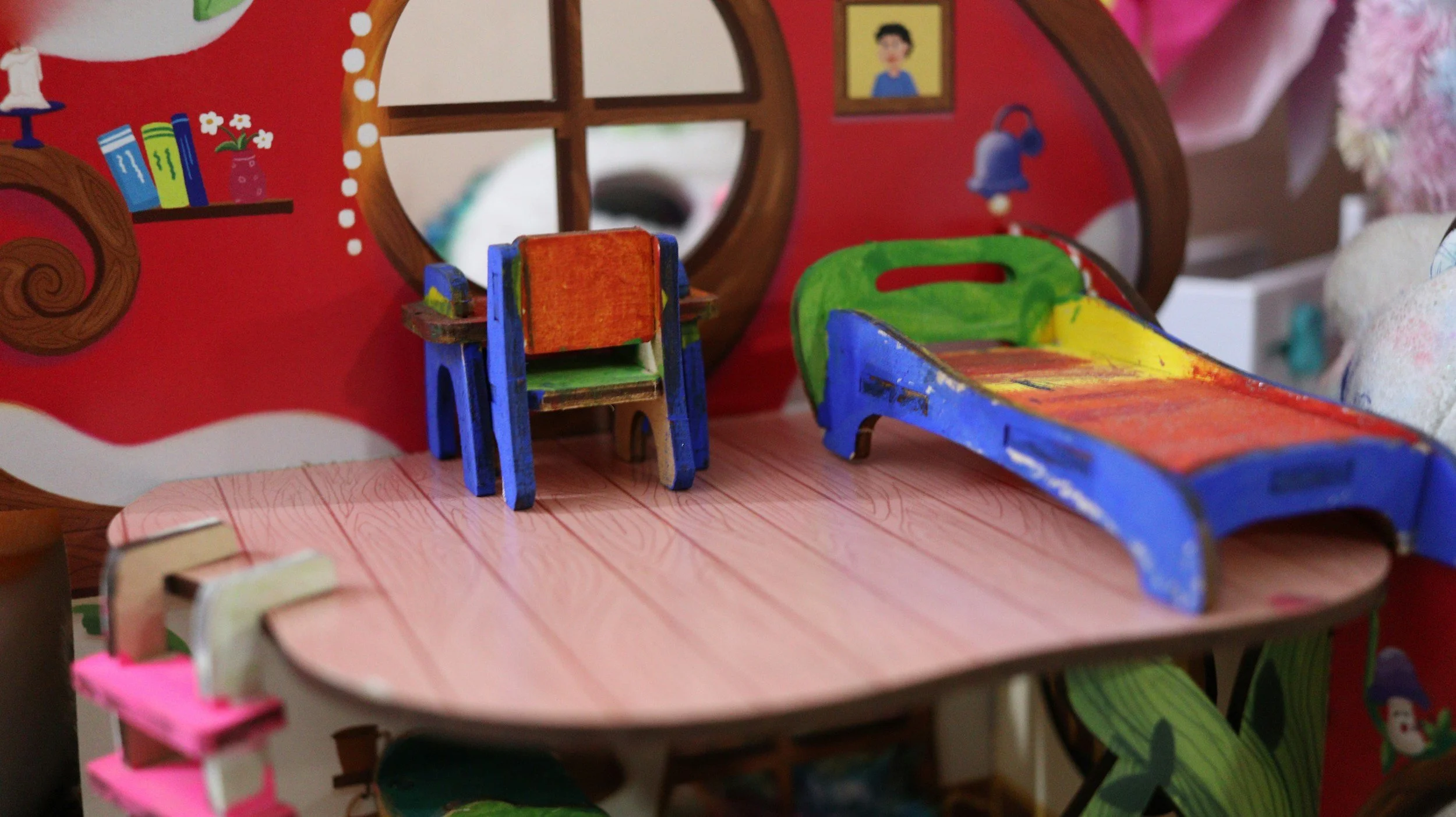 Colorful miniature wooden furniture including a small table, a chair, and a bed on a dollhouse or toy setup with decorated walls and a mirror.