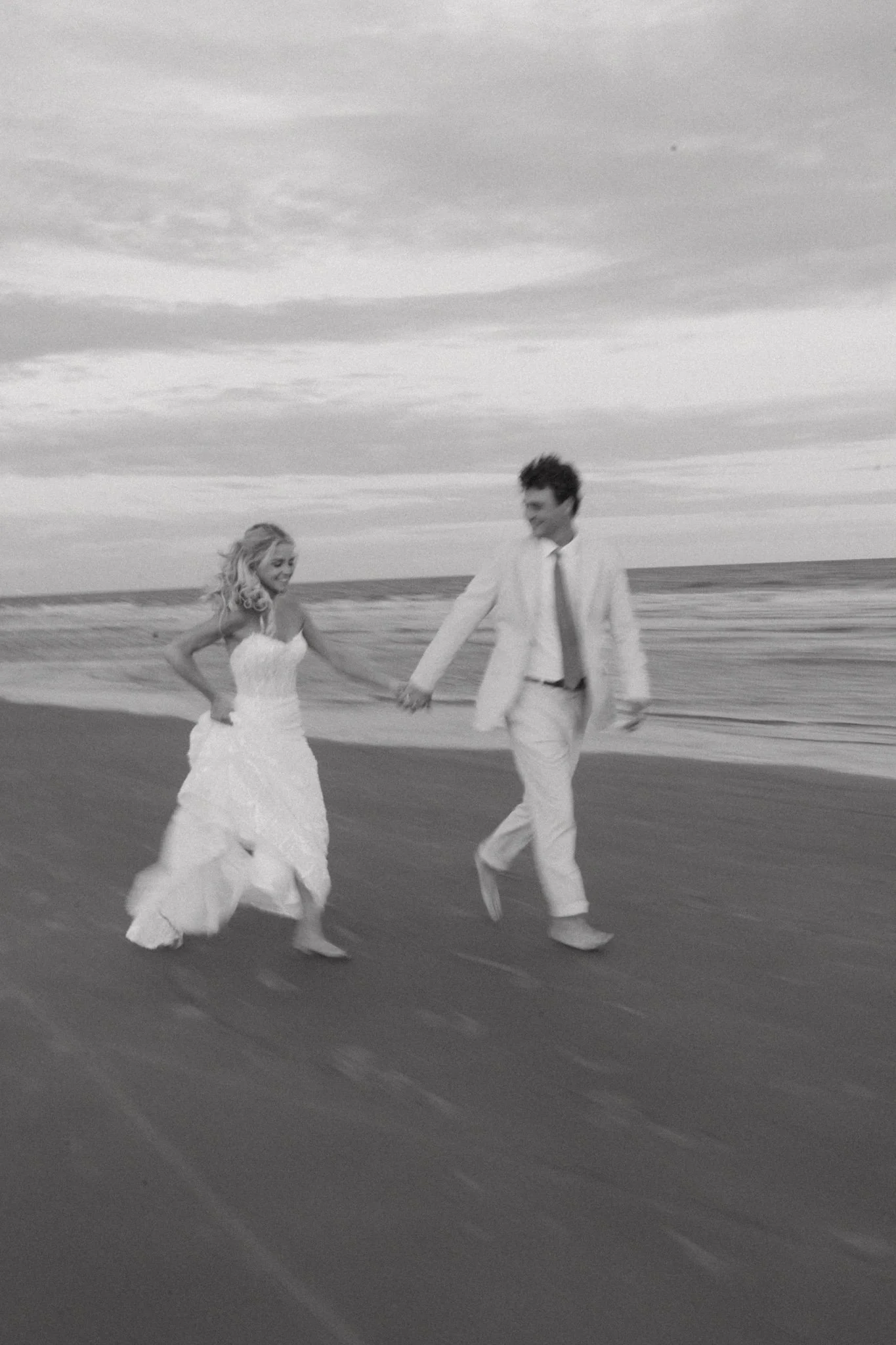 A black and white photo of a couple in wedding attire running on the beach, holding hands with a cloudy sky overhead.