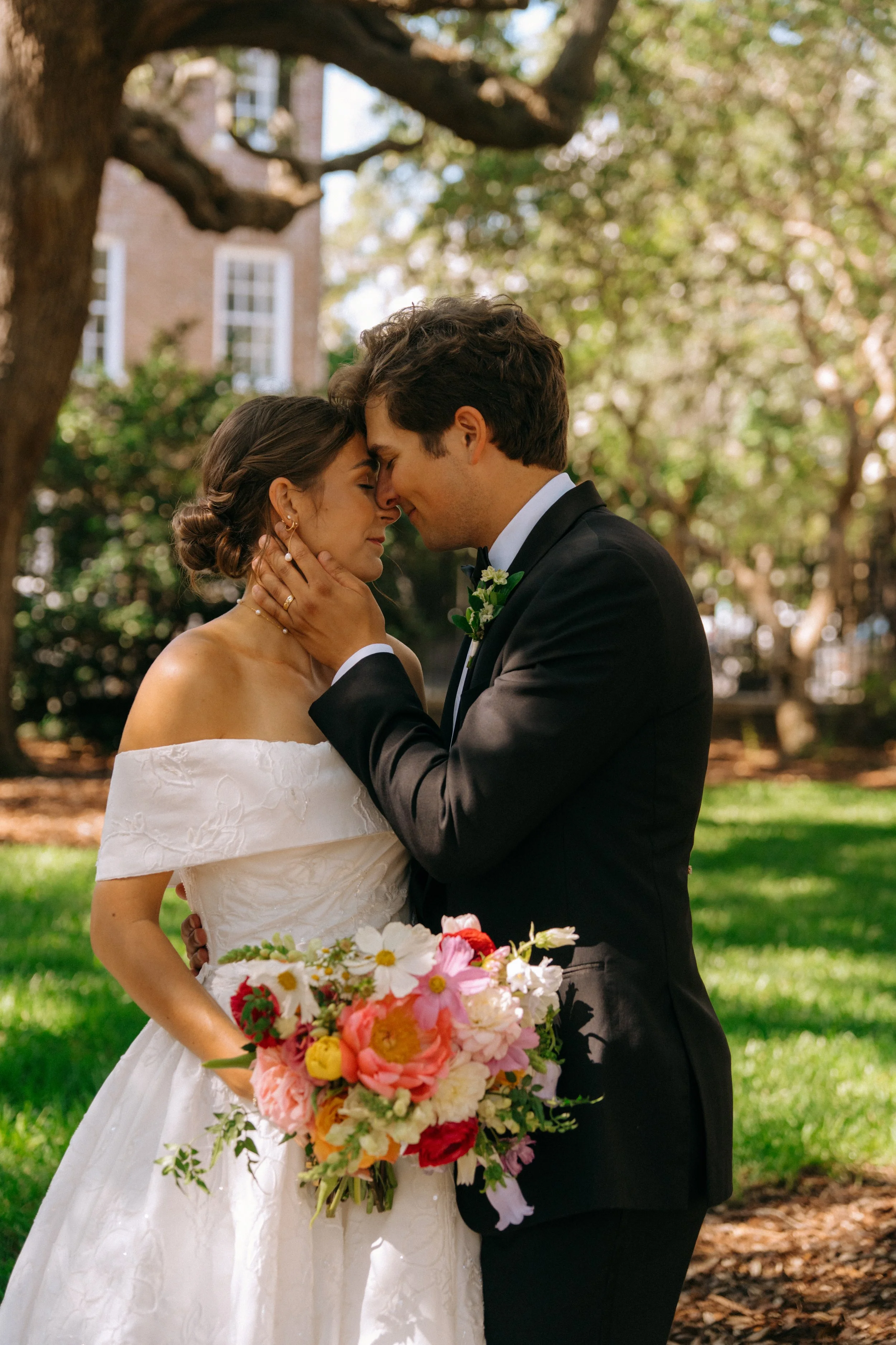 A bride and groom are in a romantic embrace outdoors, with their foreheads touching and eyes closed, holding a colorful bouquet of flowers, under a large tree with green leaves and a brick building in the background.