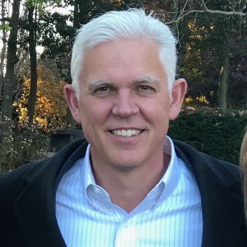 A man with white hair smiling outdoors, wearing a collared shirt and a jacket, with trees and foliage in the background.