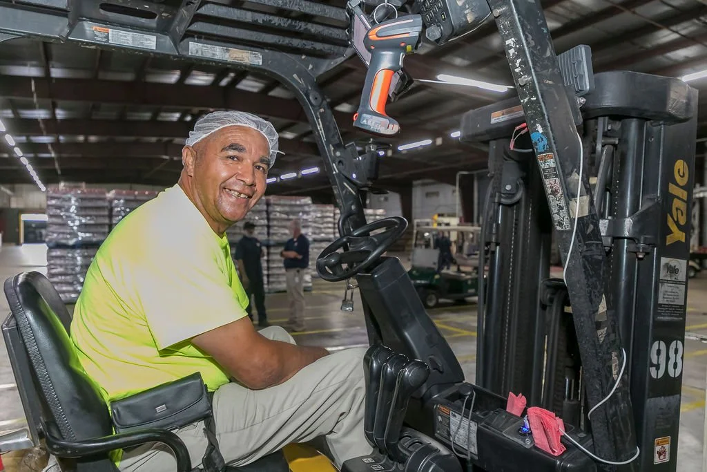A man wearing a yellow shirt and hairnet sitting on a forklift inside a warehouse.