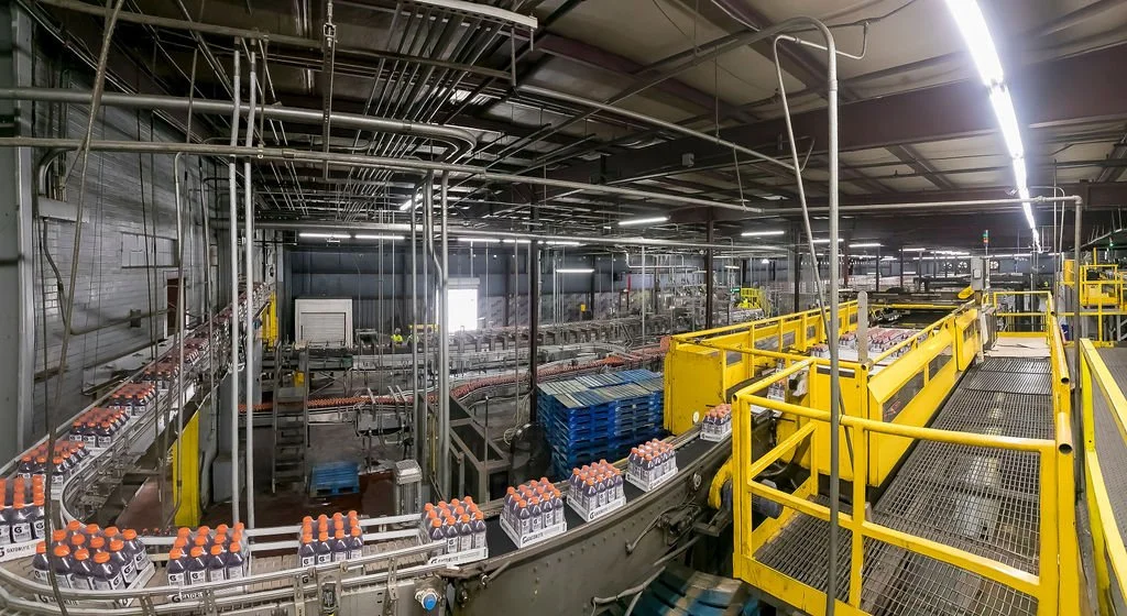 Inside a beverage bottling factory with conveyor belts carrying bottles of Gatorade, yellow safety railings, and industrial equipment.