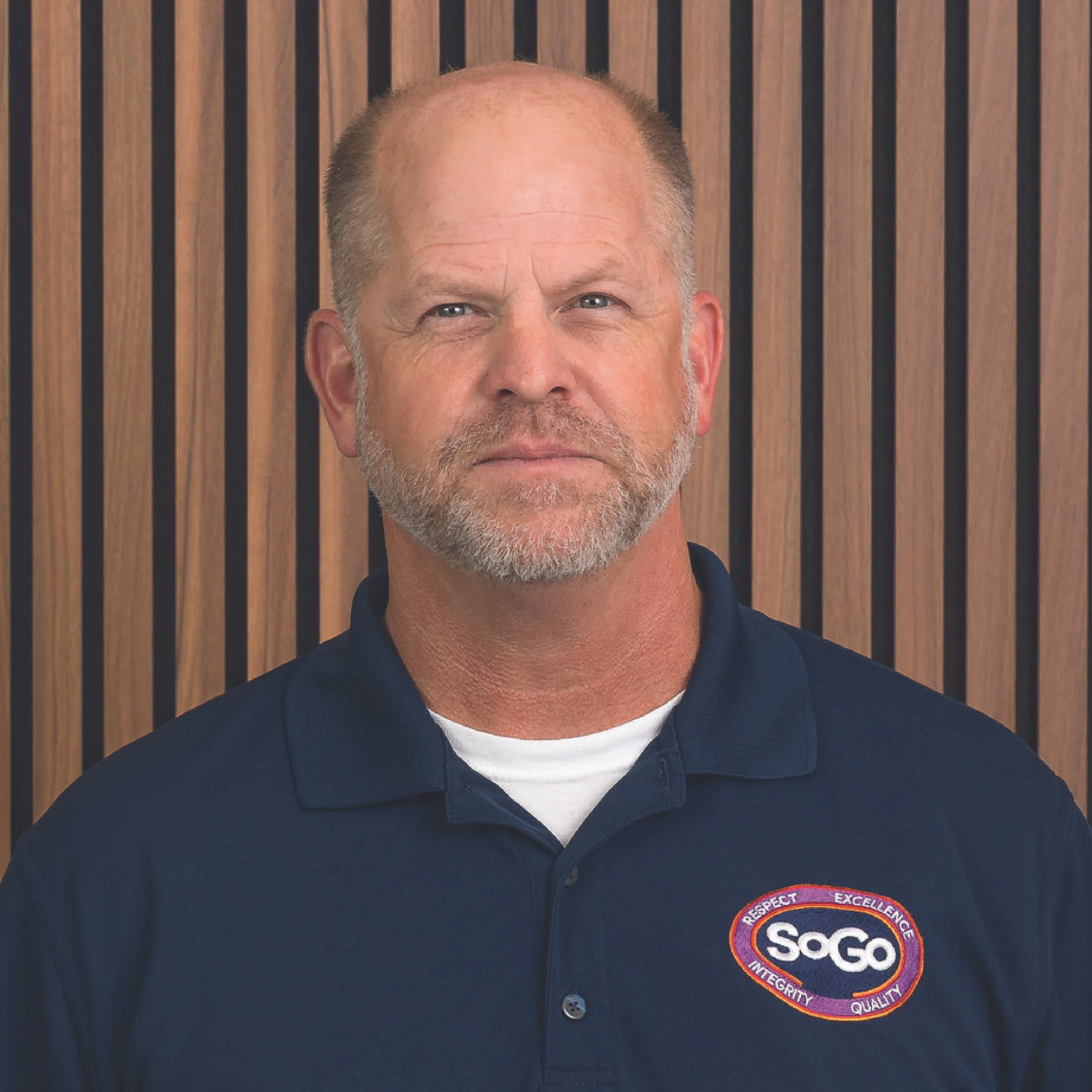 A middle-aged man with a beard and short hair, wearing a navy blue polo shirt with the SoGo logo on it, standing in front of a wooden slat wall.
