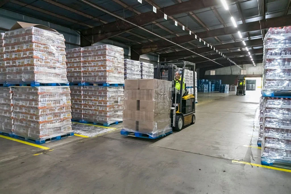 Warehouse with pallets of packaged goods, forklift operator in safety vest moving pallets, other forklifts in the background, high ceiling with industrial lighting.