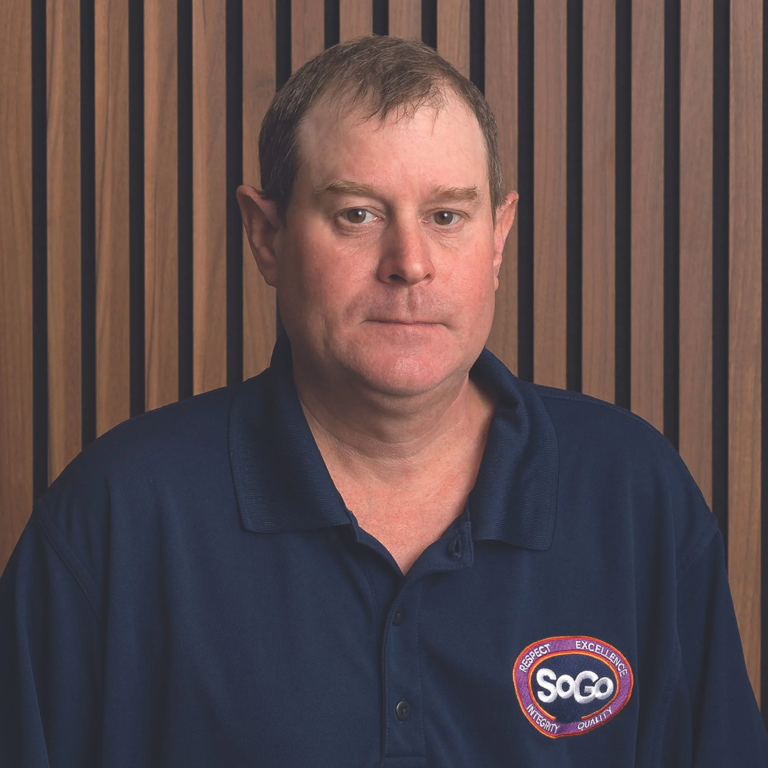 A middle-aged man with short brown hair and a serious expression, wearing a navy blue polo shirt with a SOCO logo on the chest, standing in front of a wooden panel background.