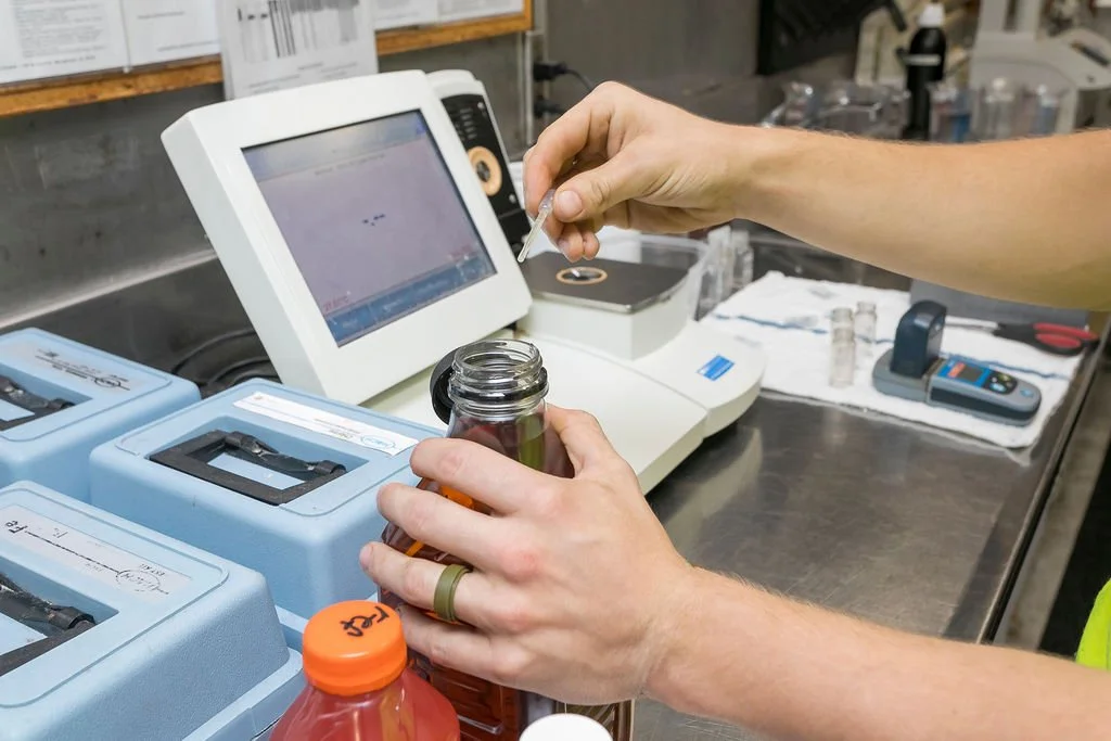 A person using a pipette to transfer liquid into a test tube on a laboratory counter, with laboratory equipment and a computer monitor nearby.