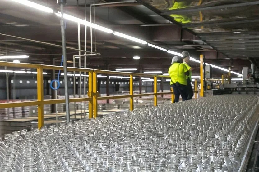 Rows of empty glass bottles on a production line in a factory with two workers in yellow safety vests and hair coverings, overseeing the process.