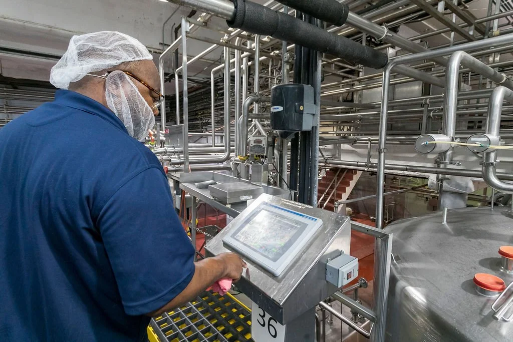 A worker in protective gear operating a control panel in an industrial facility with pipes and tanks.