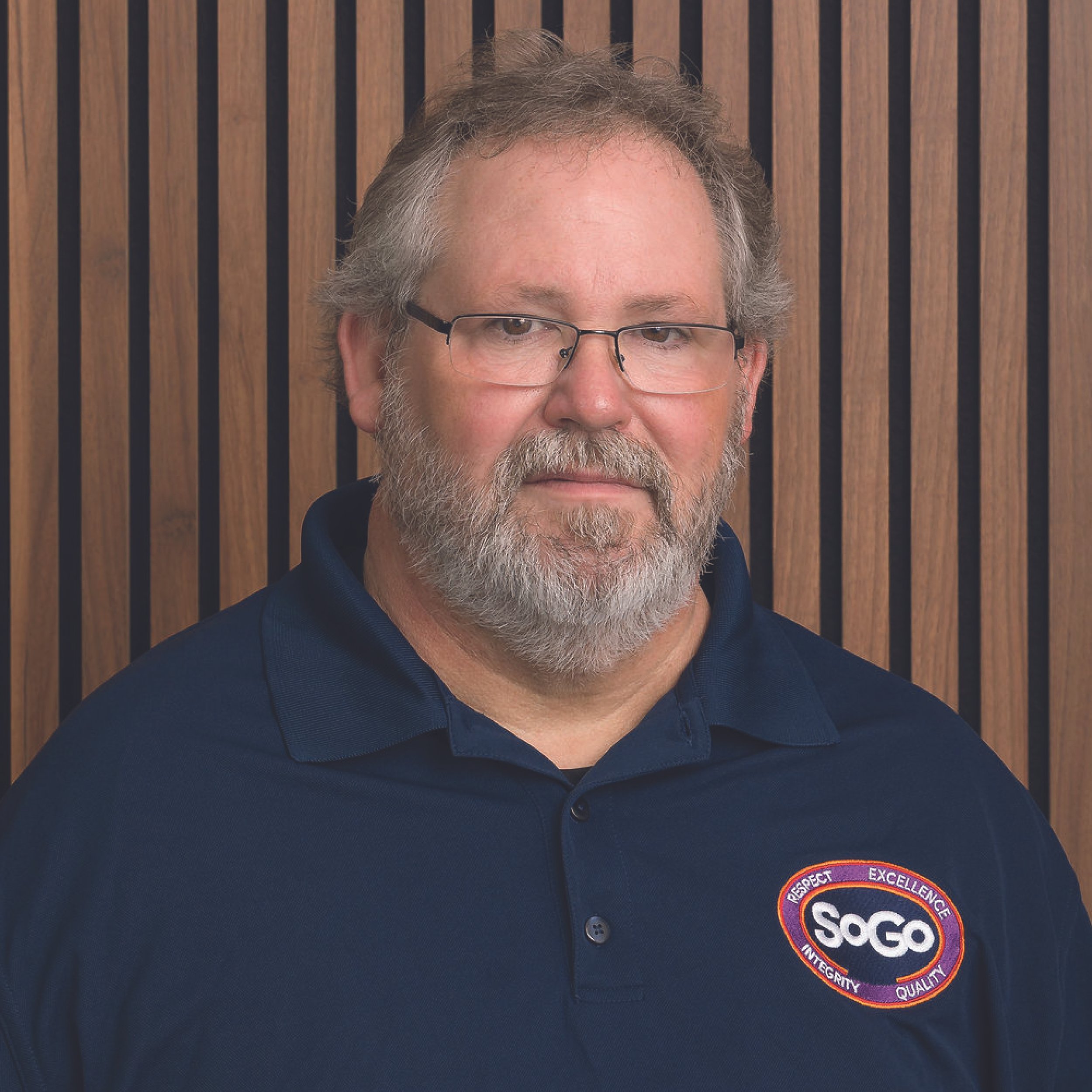 A man with glasses, a beard, and gray hair, wearing a navy blue polo shirt with a logo on the chest that reads 'SoGo' and words 'Respect, Excellence, Integrity, Quality' around it, standing in front of a wooden slat wall.