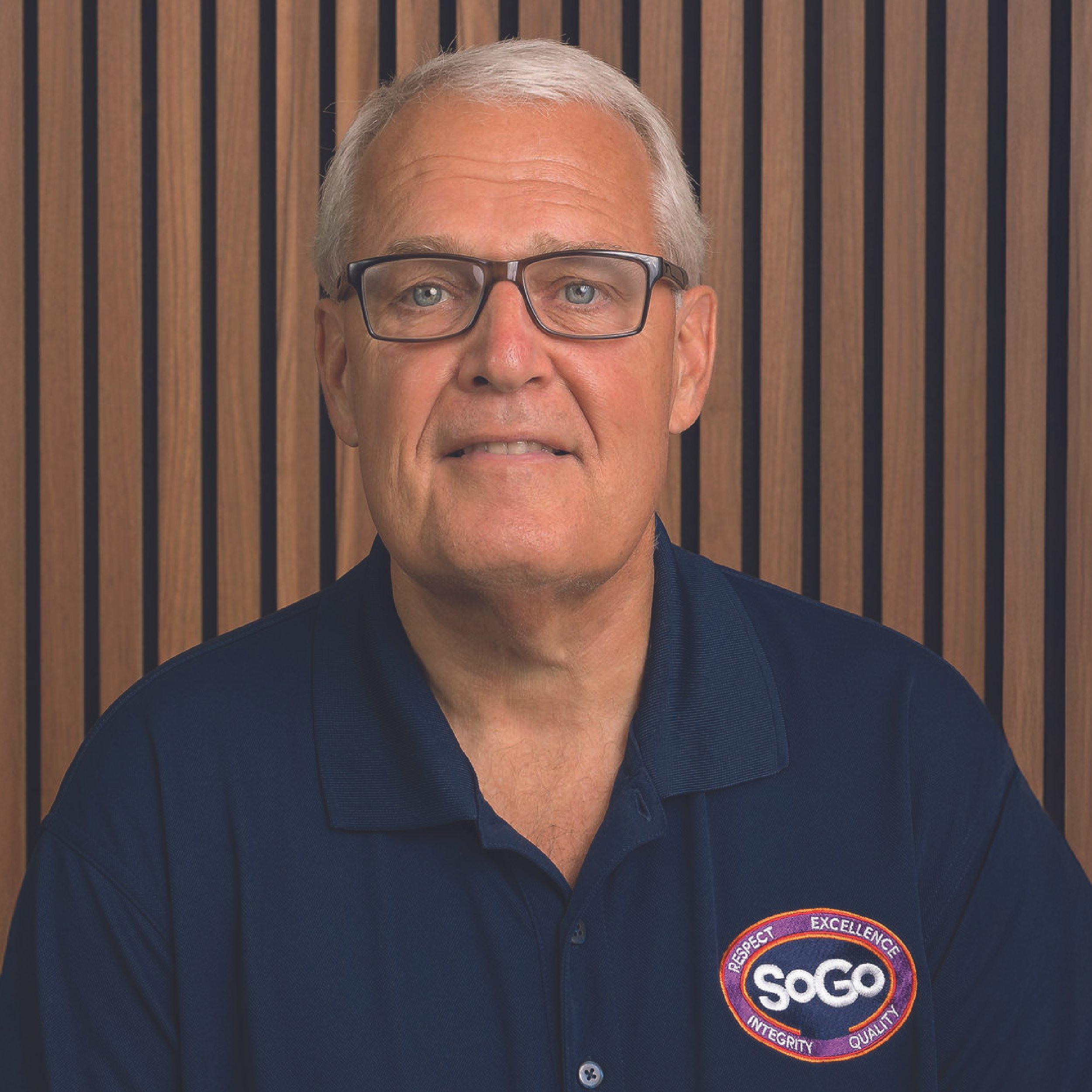 A man with gray hair and glasses wearing a navy polo shirt with a SOGO logo, sitting in front of a wooden panel background.