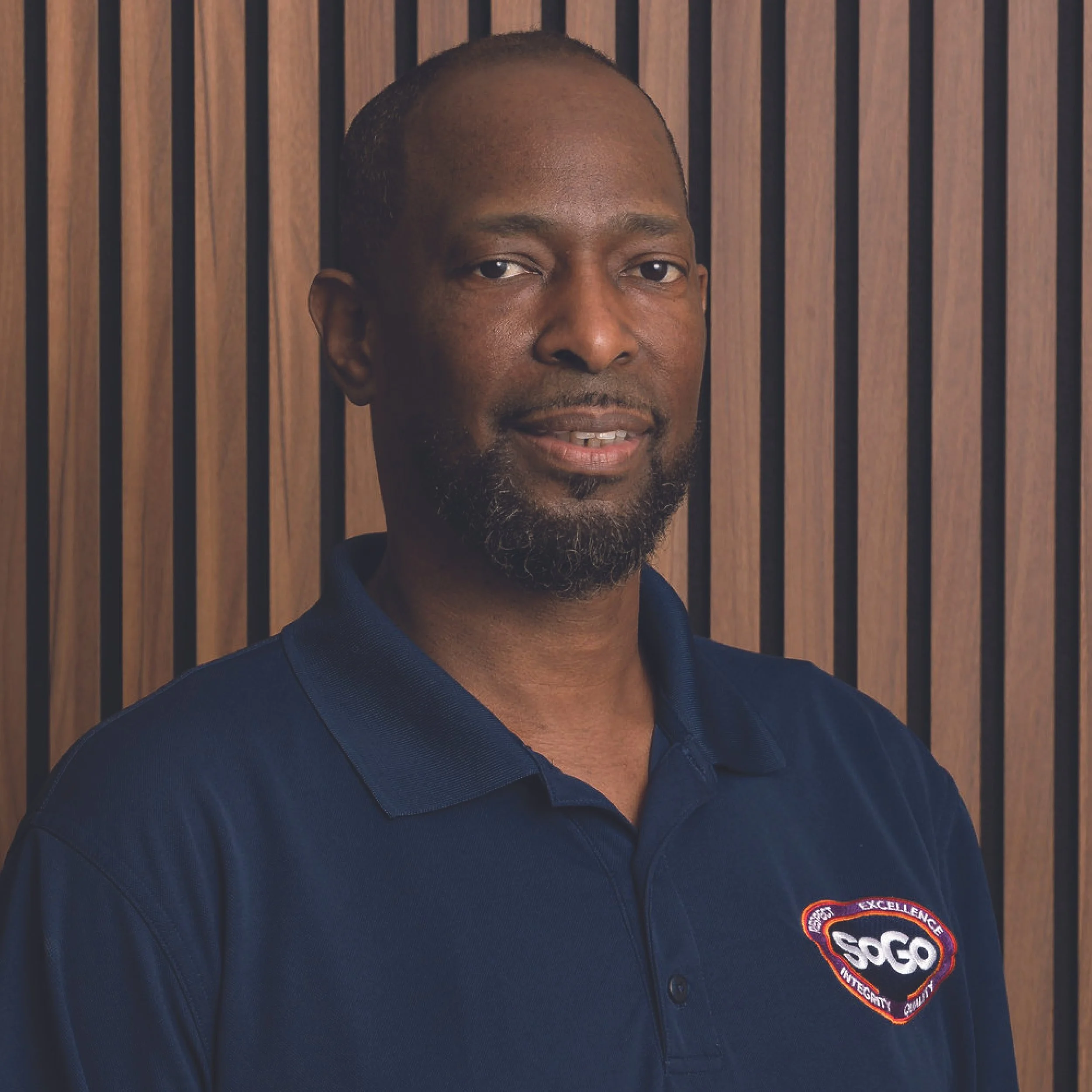 Portrait of a man with a beard wearing a navy blue polo shirt with a logo that reads 'SoGo' standing in front of a wooden panel wall.
