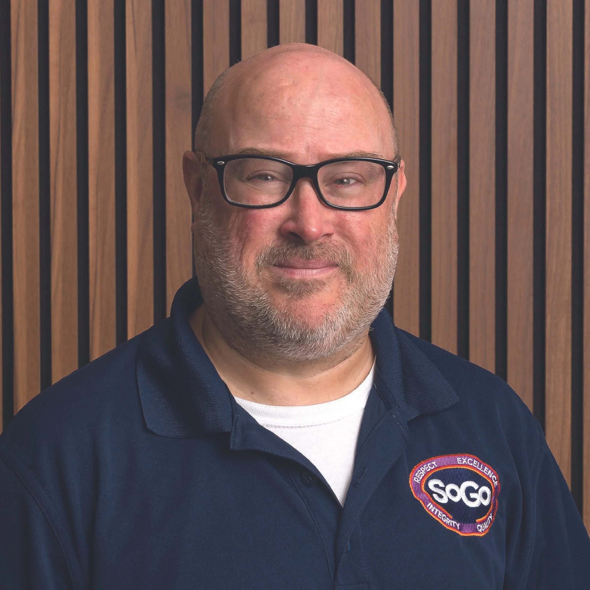A man with glasses and a beard wearing a navy blue uniform with a SAGO logo in front of a wooden panel background.