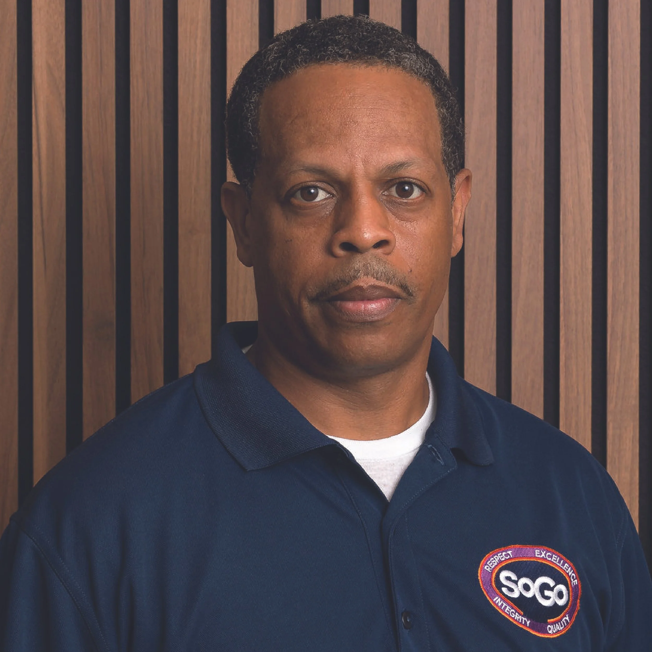 A man with short curly hair wearing a navy blue polo shirt with a SOGO logo and a wooden paneled background.