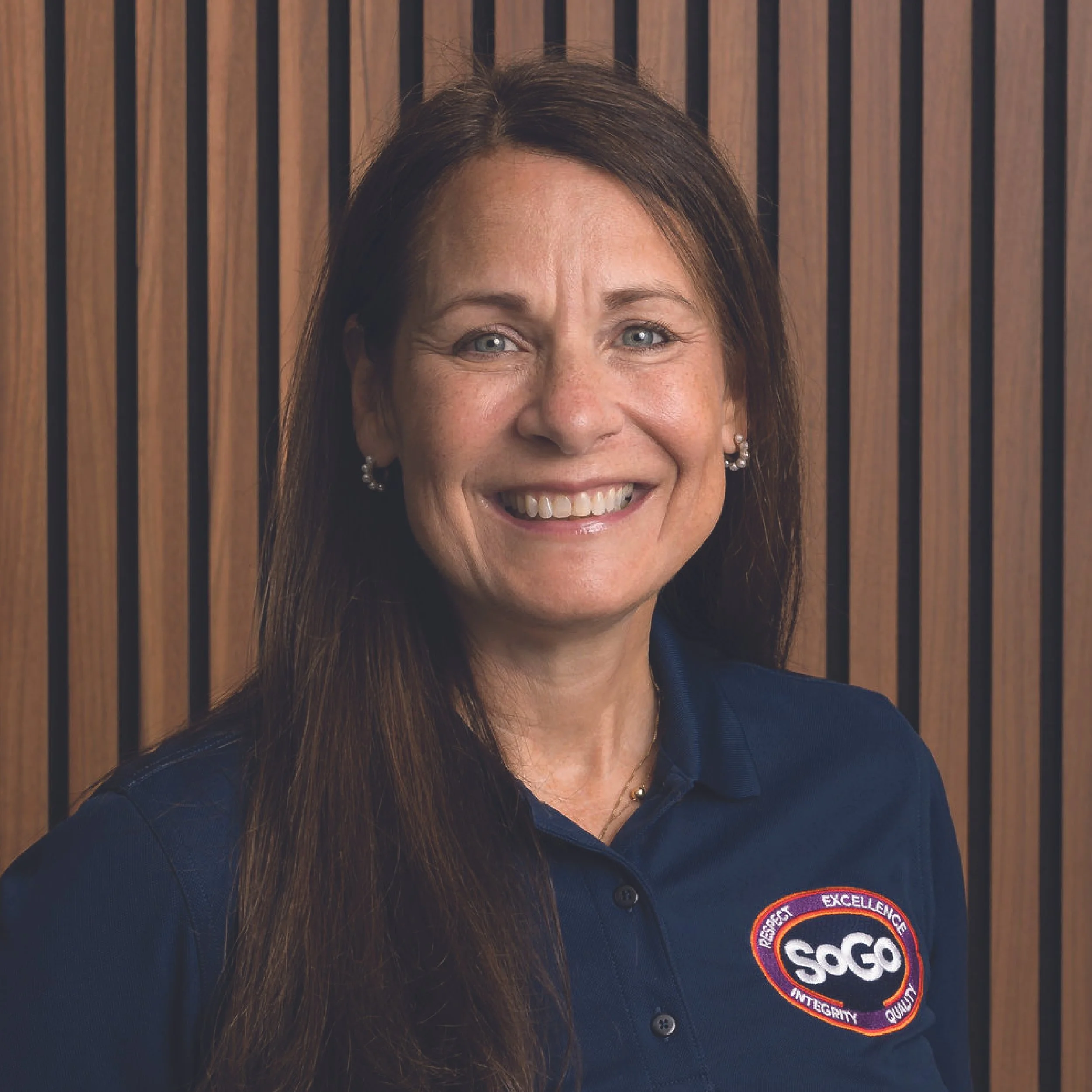 A woman with long brown hair smiling, wearing a navy blue polo shirt with a logo that says SOGO, against a wooden background.