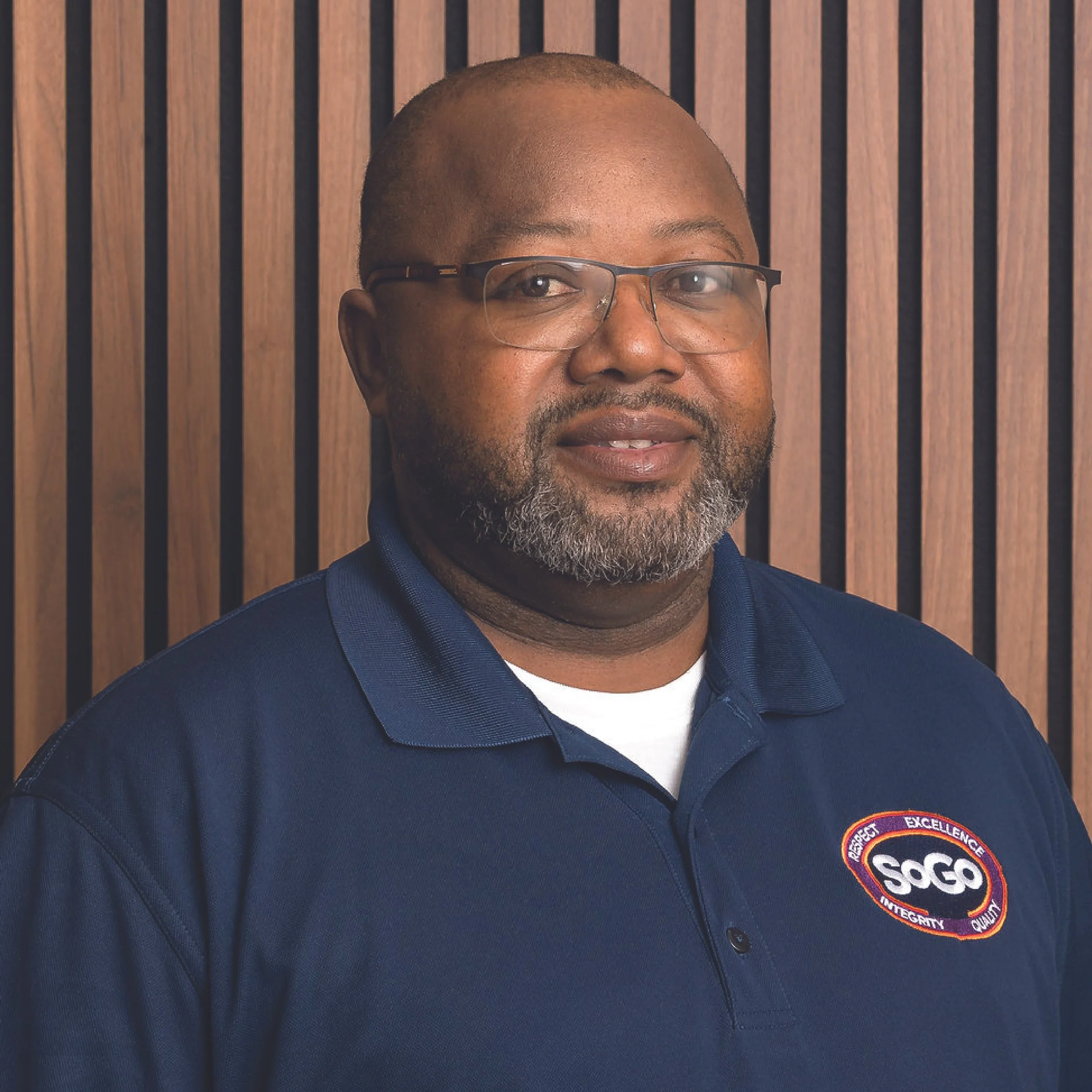 A man with glasses and a beard is wearing a navy polo shirt with a SOG logo, standing in front of a wooden slat wall.