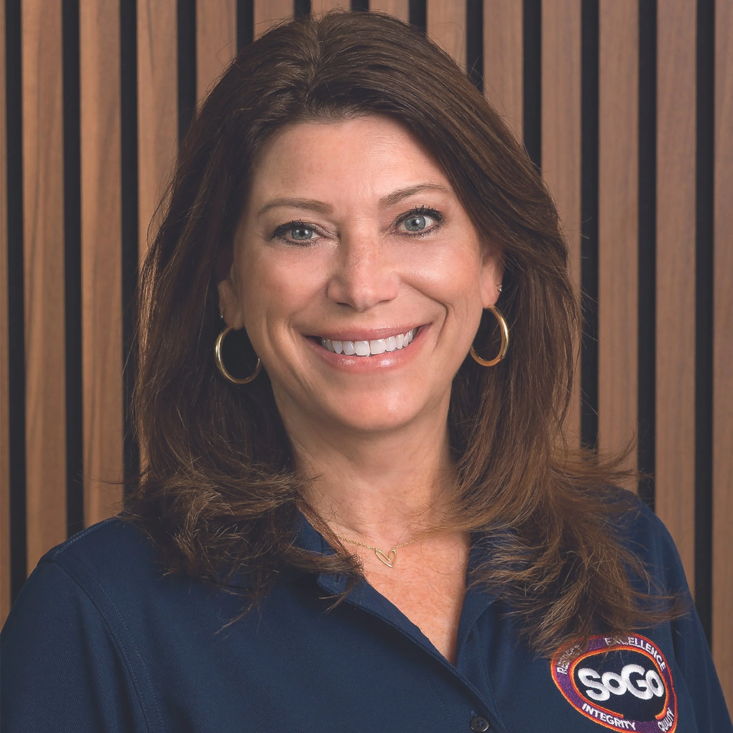 A smiling woman with shoulder-length brown hair, wearing hoop earrings and a navy blue polo shirt with a SOG logo, standing in front of a wooden panel background.
