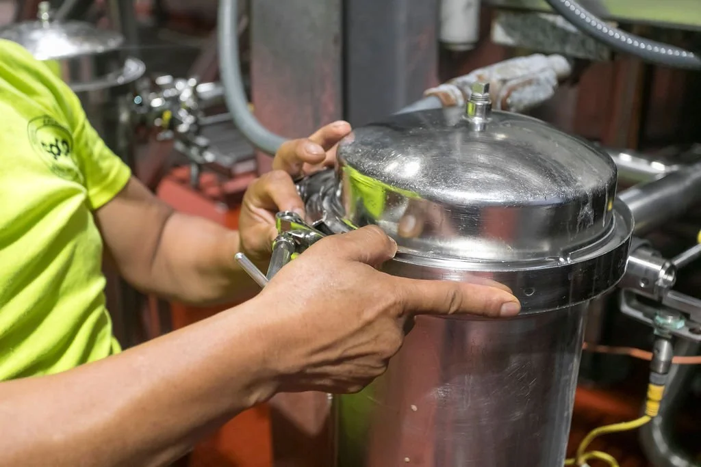 A person wearing a neon green shirt is adjusting a shiny metal tank fitted with a clear plastic lid, in a mechanical or industrial setting.