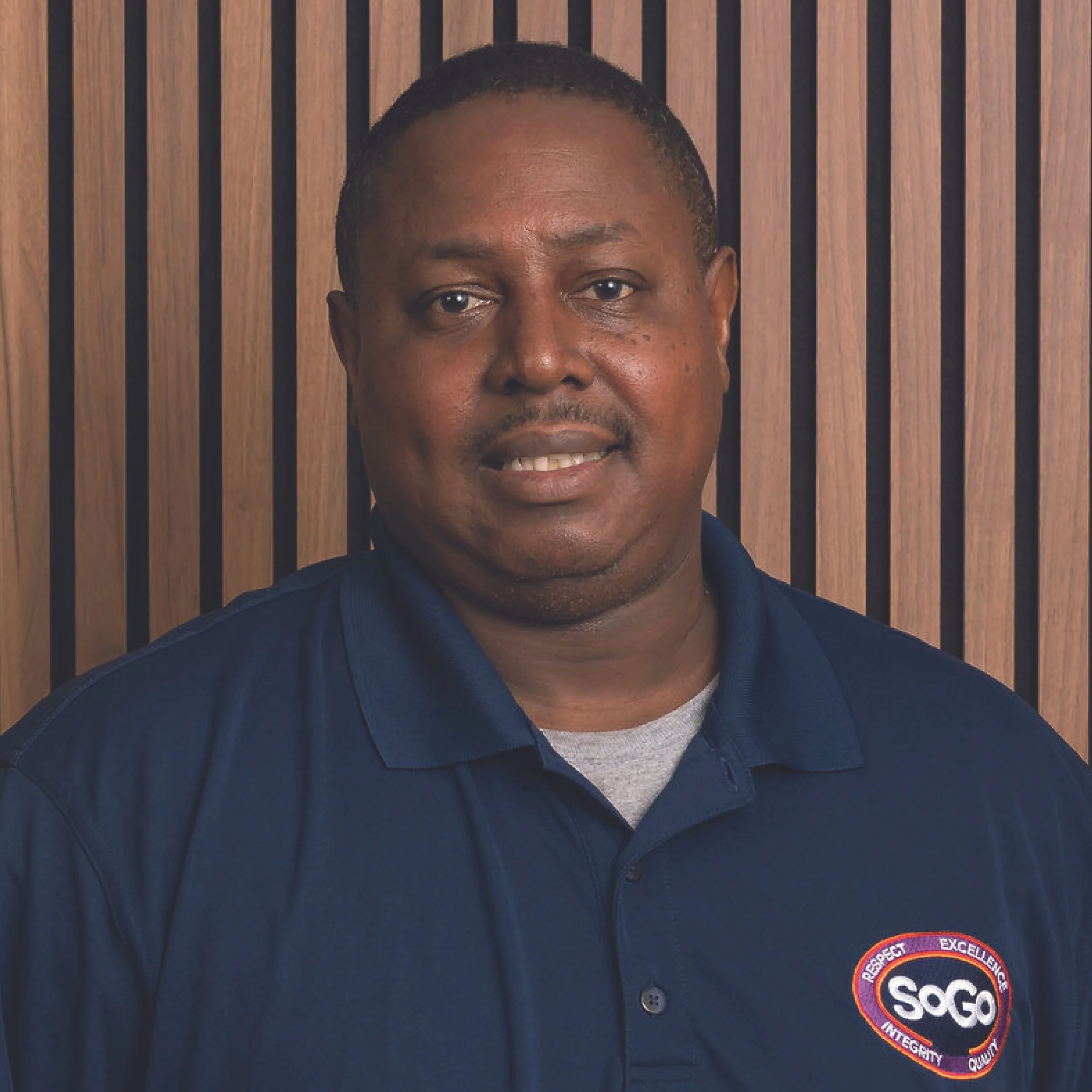 A man wearing a navy blue polo shirt with a logo on the chest, standing in front of a wooden panel background.