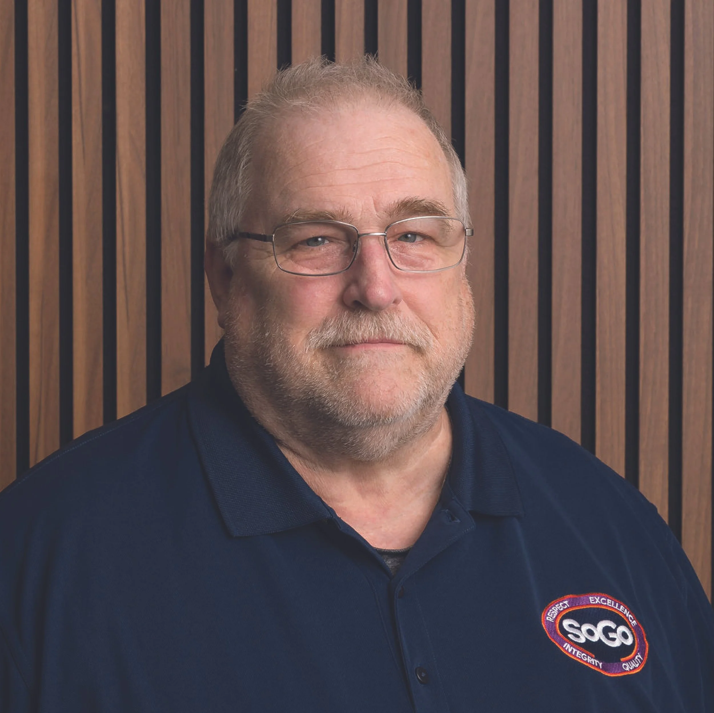 Headshot of a middle-aged man with glasses and a beard, wearing a navy blue collared shirt with SOGO logo, standing in front of a wooden slat background.