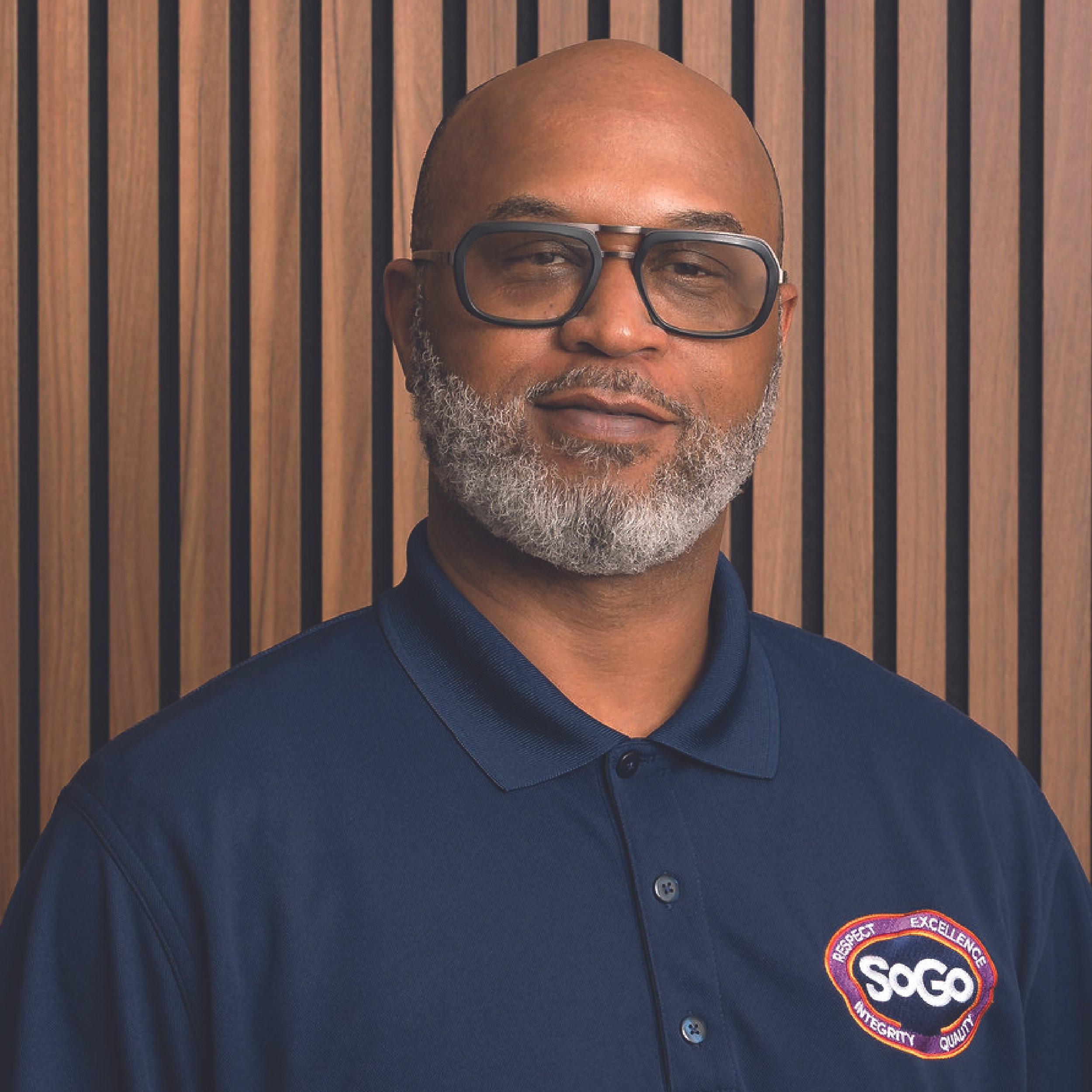 A man with glasses and a gray beard wearing a navy blue polo shirt with a SOGO logo, standing in front of a wooden slat wall.