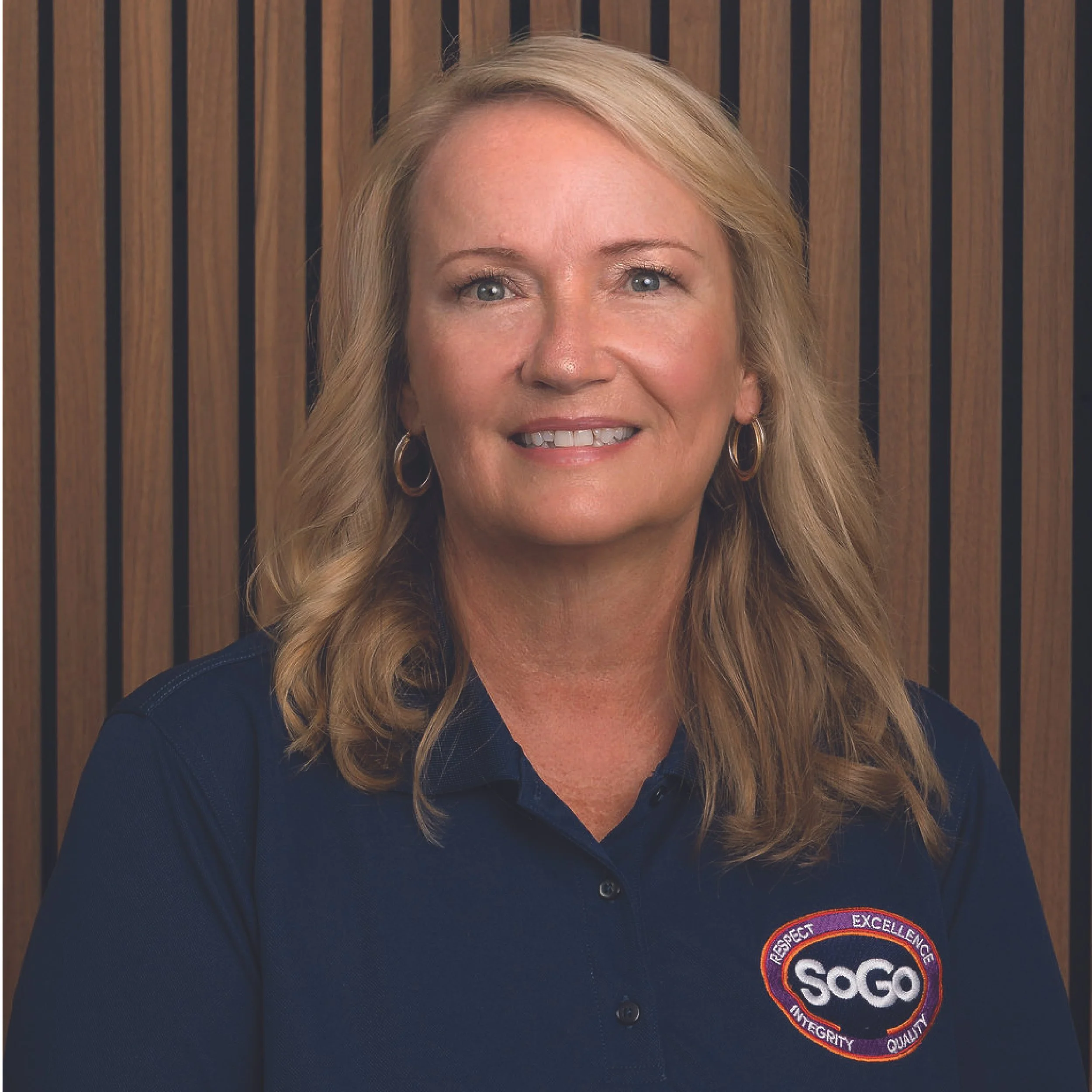 A woman with blonde hair smiling, wearing a navy polo shirt with a SOGO logo patch that includes the words Respect, Excellence, Integrity, and Quality, with a wooden panel background.