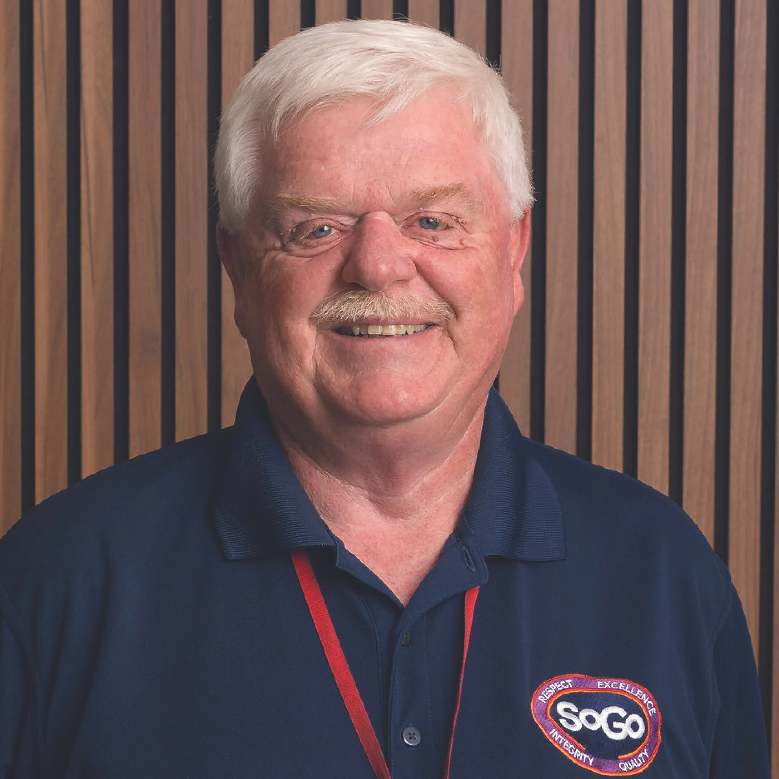A smiling elderly man with white hair and a mustache wearing a navy blue polo shirt with a SOGO logo and a lanyard, standing against a wooden panel background.