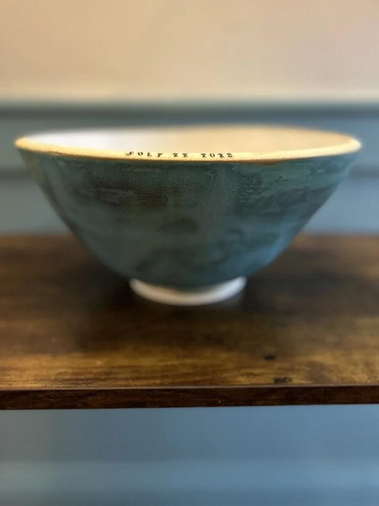 Close-up of a ceramic bowl with a green exterior and white rim, placed on a wooden surface. The bowl has printed text around the rim indicating a date, 'JULY 22 2018'.