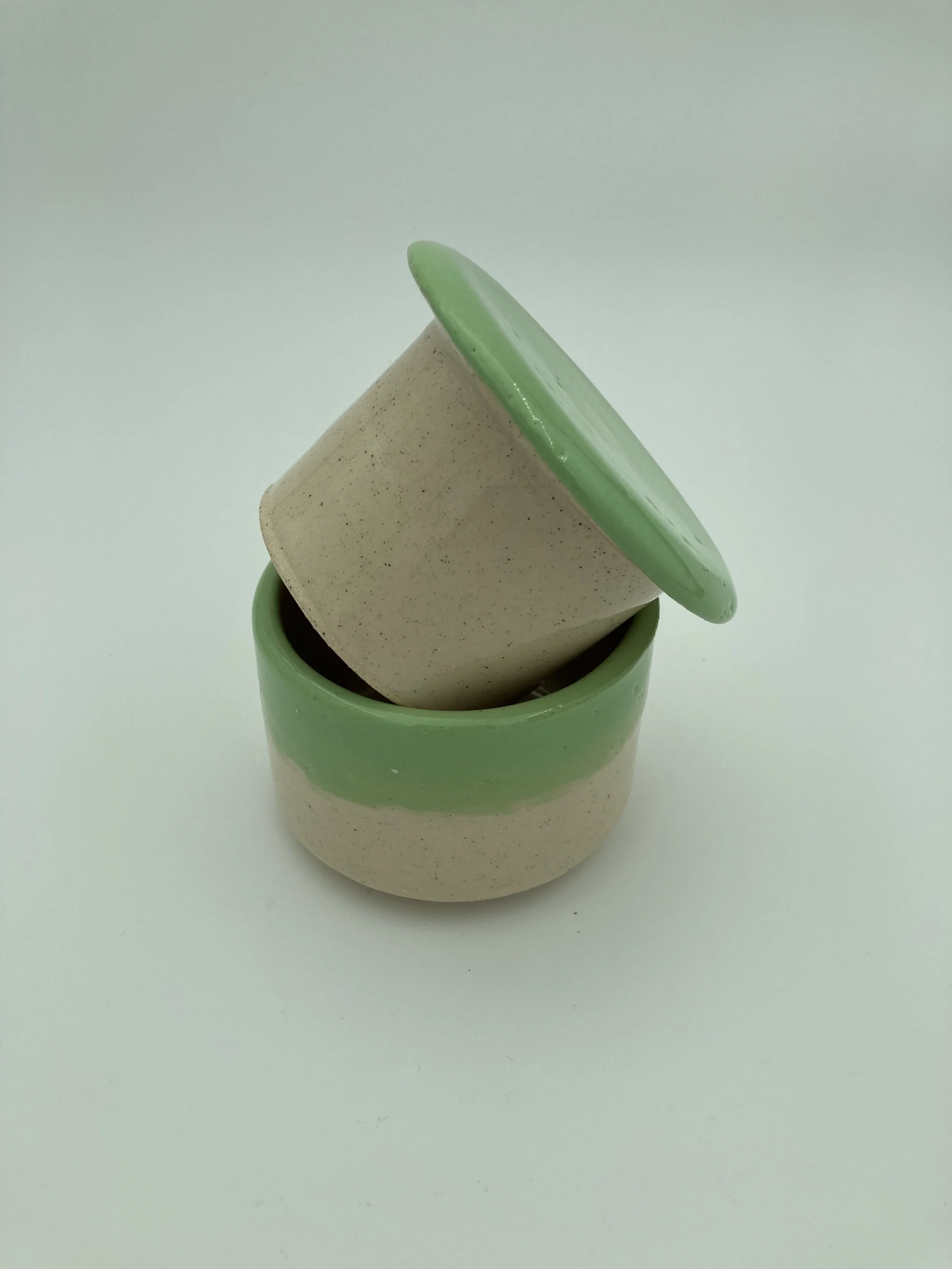 Two stacked ceramic plant pots, one beige and one with a green top rim, on a plain white background.