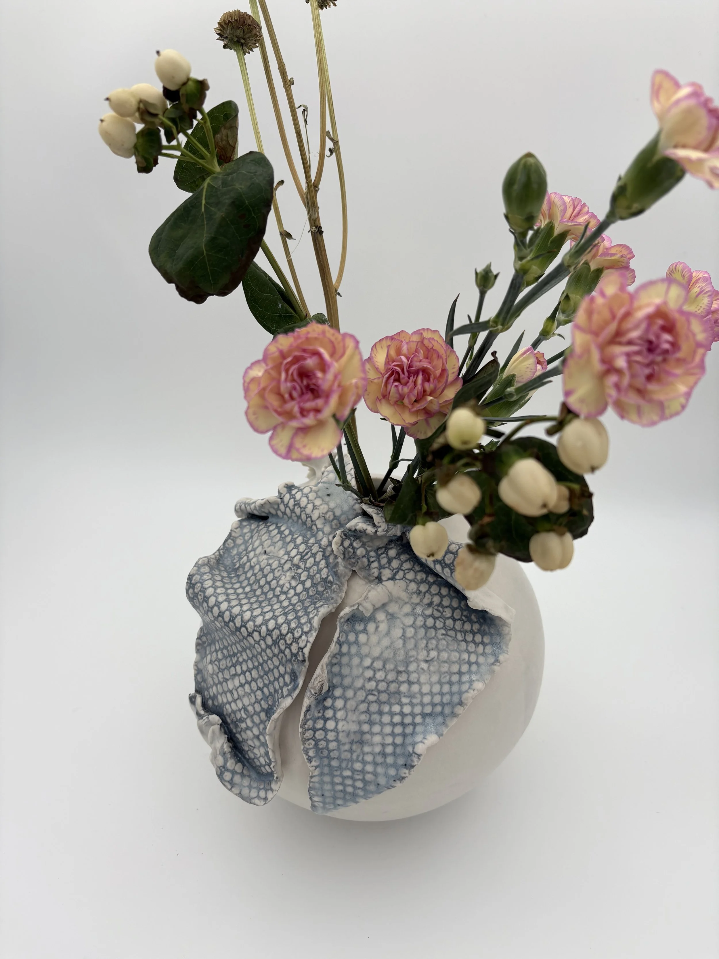 A white vase with a textured surface holds a bouquet of pink and cream flowers with green leaves and stems, including some small white buds, set against a plain white background.