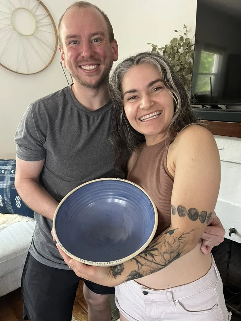 A man and a woman smiling and standing close together indoors. The woman is holding a large, empty blue bowl. She has gray hair, tattoos on her arm, and is wearing a sleeveless brown top and white pants. The man has a receding hairline, is wearing a