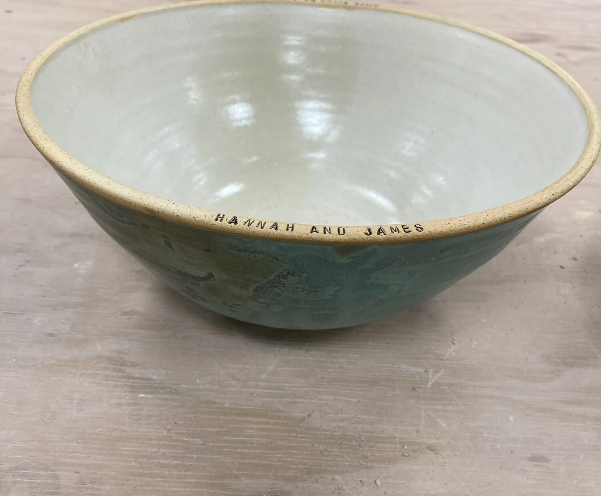 A ceramic bowl with a glazed interior and a matte, speckled exterior. The rim has an inscription that reads 'HANNAH AND JAMES.' The bowl is placed on a light wooden surface.