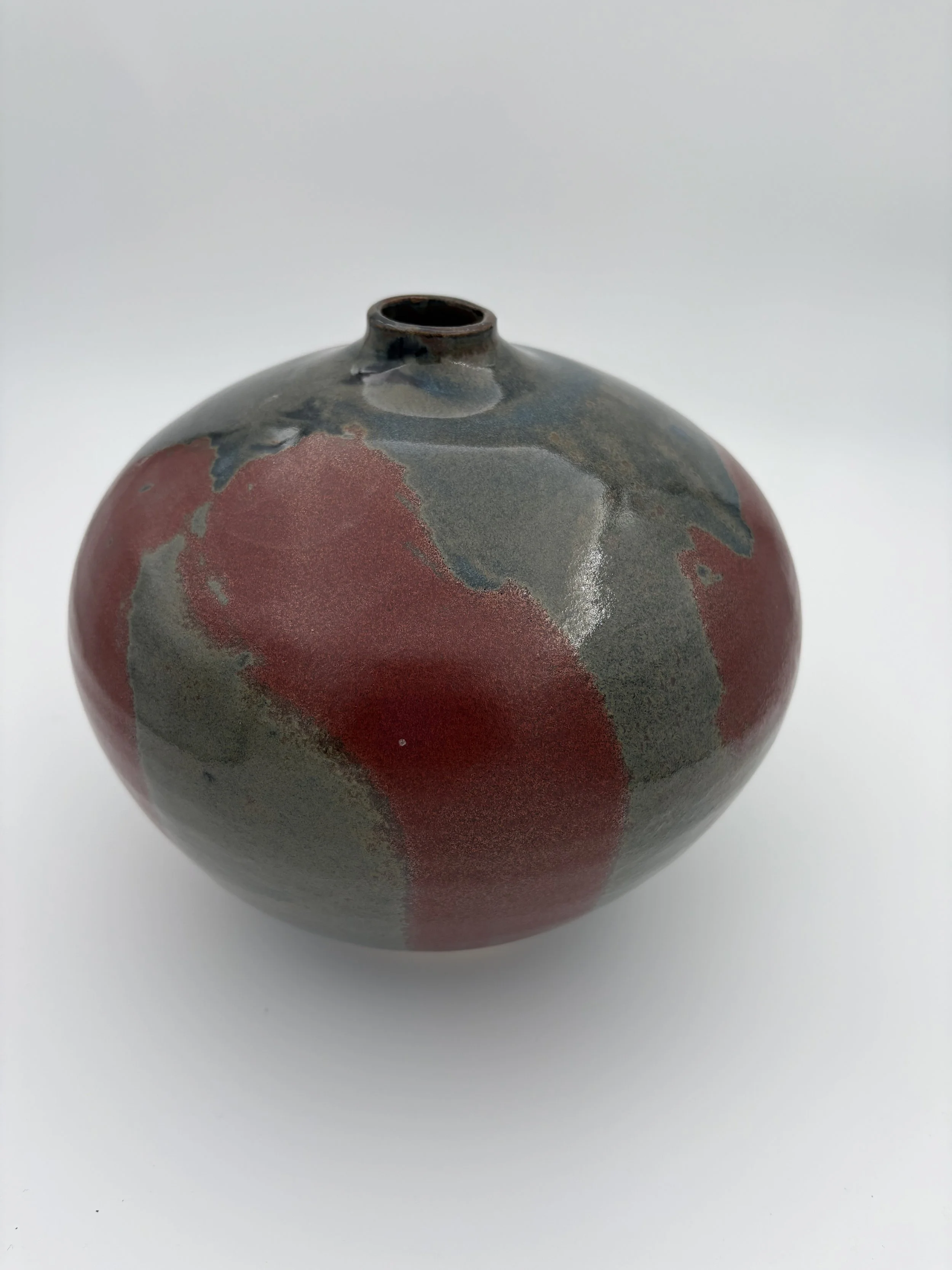 A round ceramic vase with a narrow opening, decorated with abstract red, gray, and black glaze patterns.