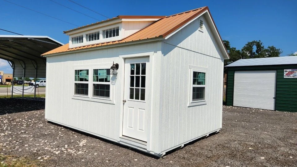 Small white shed with a gabled roof and multiple windows, situated outdoors on a gravel lot.