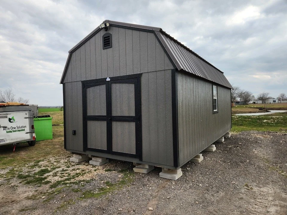 A gray metal shed on cinder blocks in an open area with a cloudy sky.
