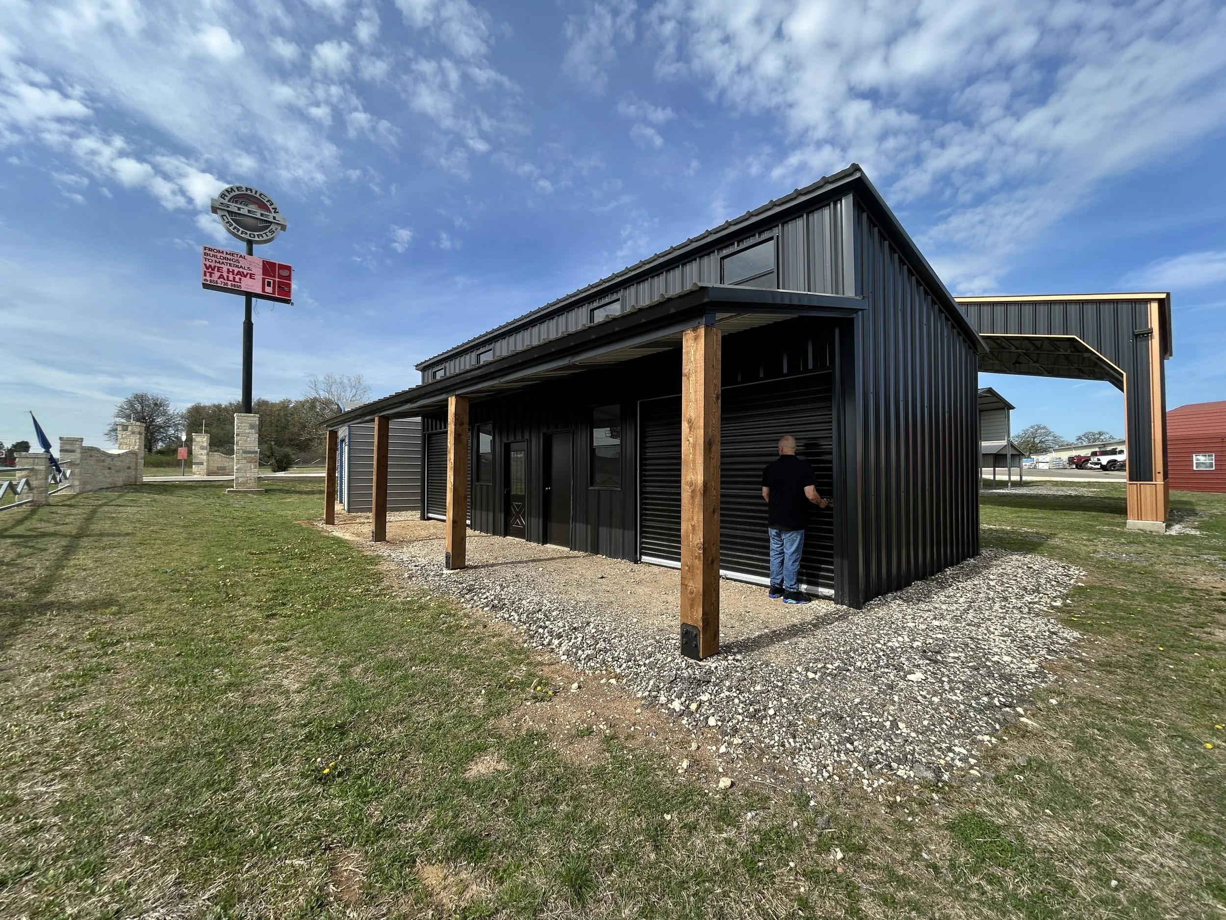 A black metal building with wooden support beams and a sloped roof, situated on a gravel and grassy lot under a blue sky with scattered clouds.