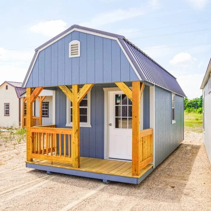 Small blue house with wooden porch and railing, white door, and metal roof, situated outdoors on a dirt lot.
