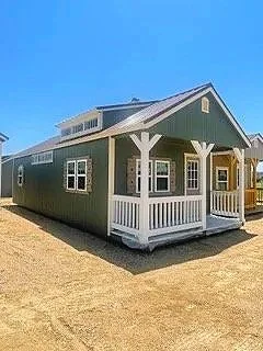 A large, gray cabin with white trim and a porch, set on a dirt lot under a clear blue sky.