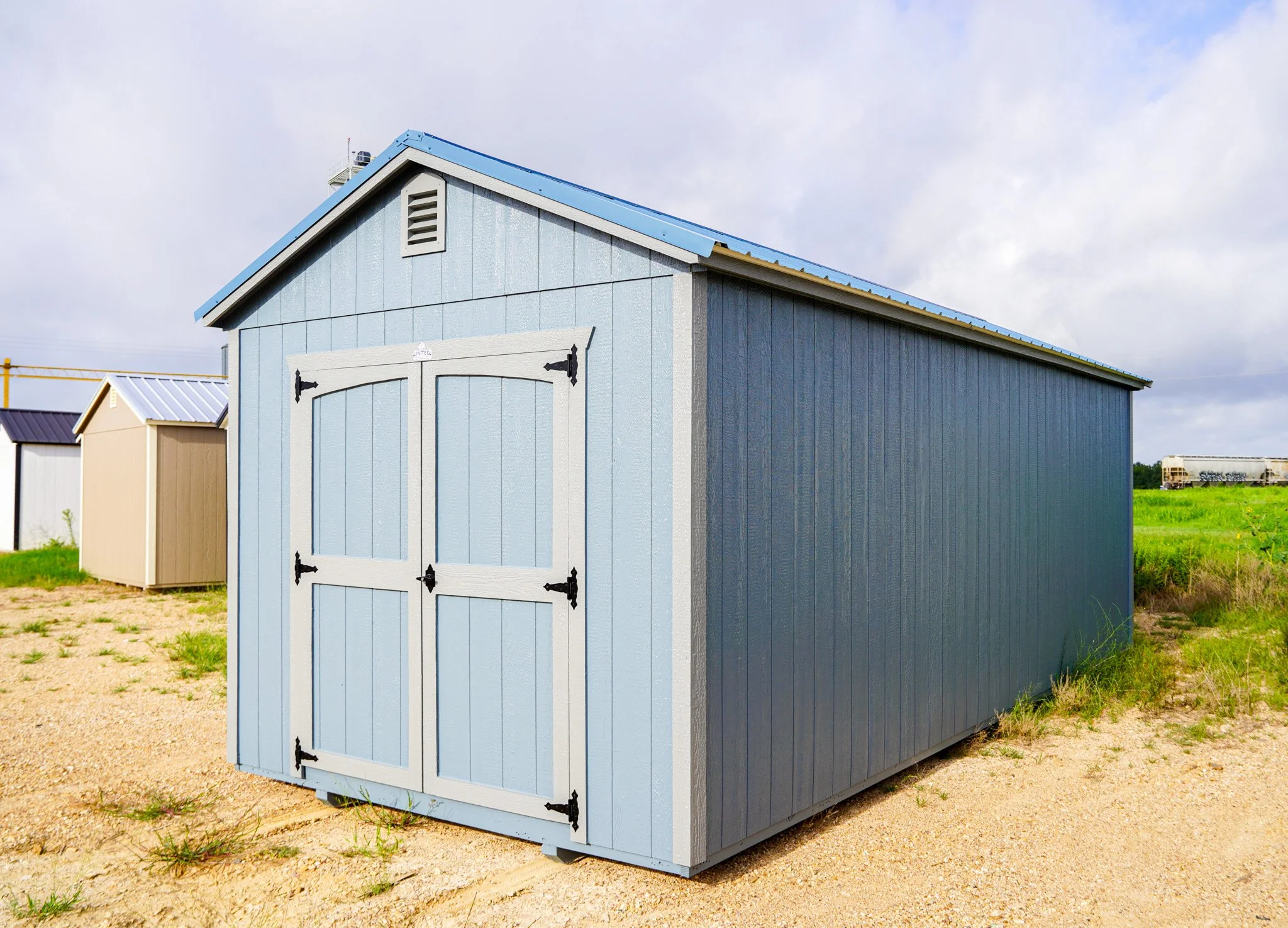 Blue storage shed with white double barn doors, set on a gravel lot with grass under a cloudy sky.