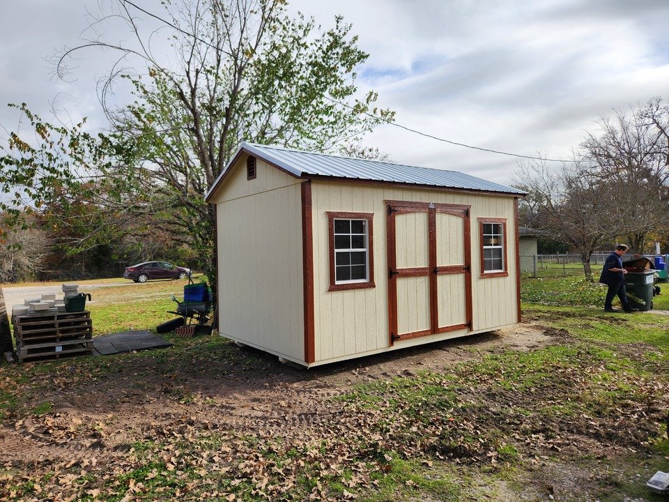 A small beige shed with brown trim and double doors, situated on a patch of dirt and grass, with two small windows and a sloped metal roof, in a backyard with trees.