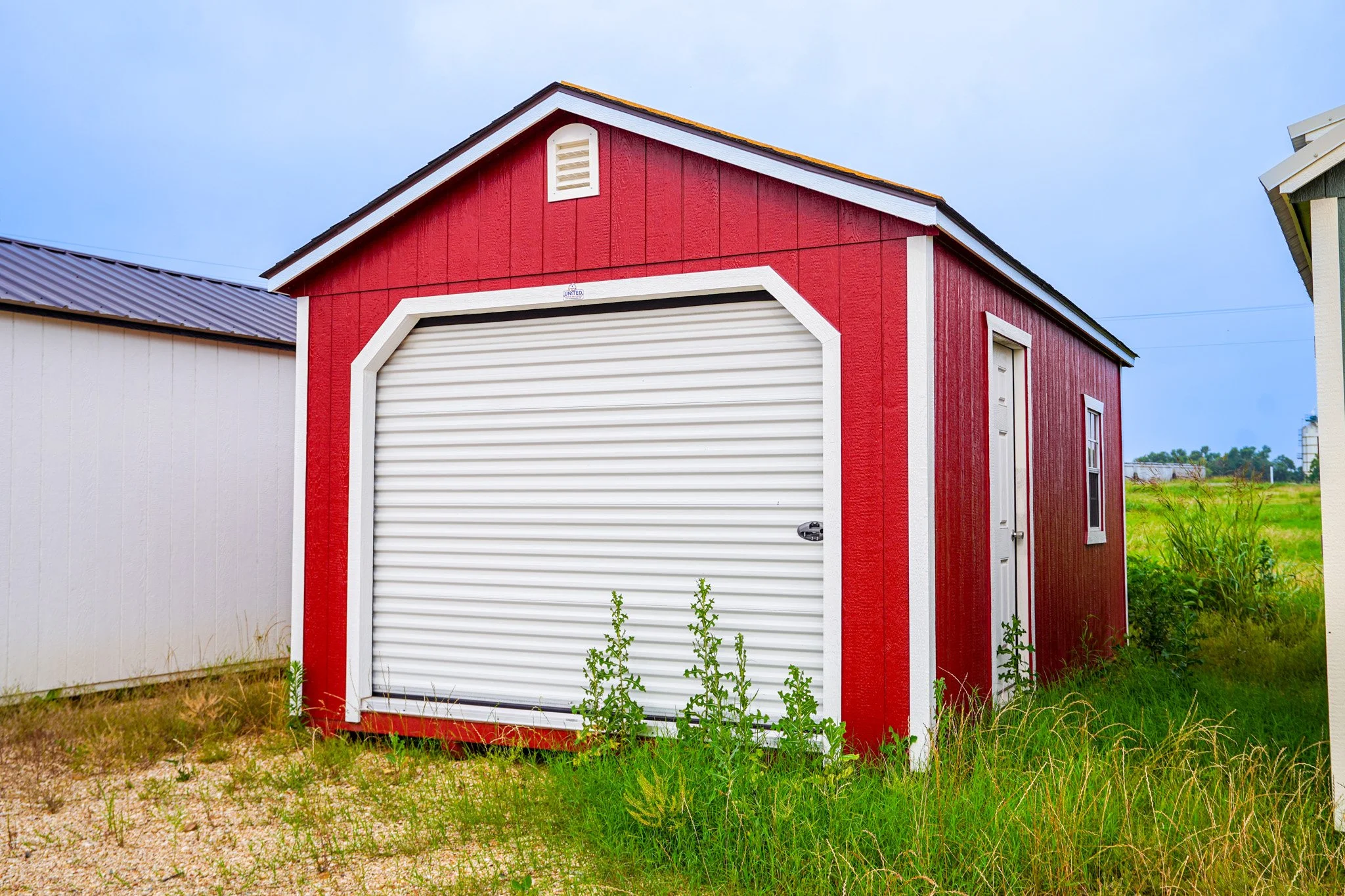 A red storage shed with white trim, featuring a large roll-up door and a side door, situated on grass and dirt, with other buildings in the background under a cloudy sky.