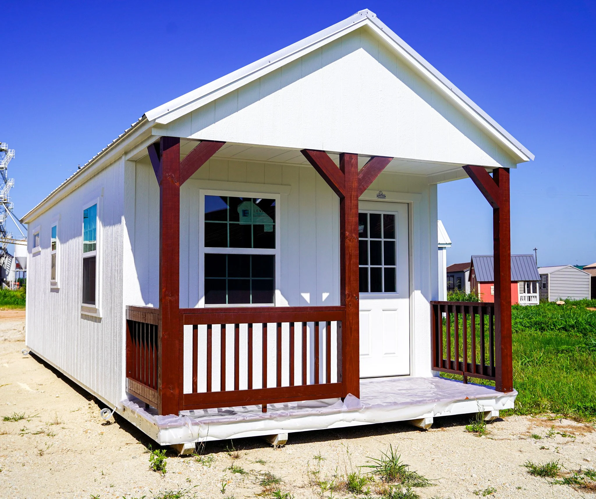 A small white prefabricated cabin with a front porch, wooden railing, and a gabled roof, situated on a sandy lot with a grassy area nearby.