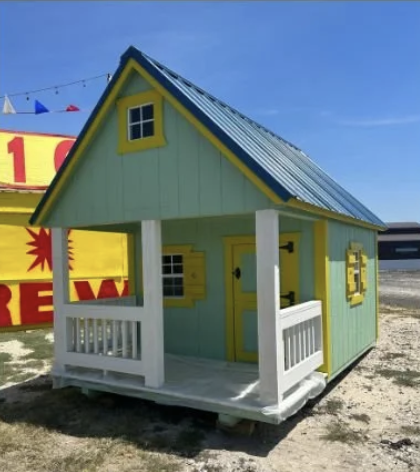 Small mint green playhouse with yellow trim, a small porch with white railing, and a slanted metal roof, set outdoors on a gravel surface.