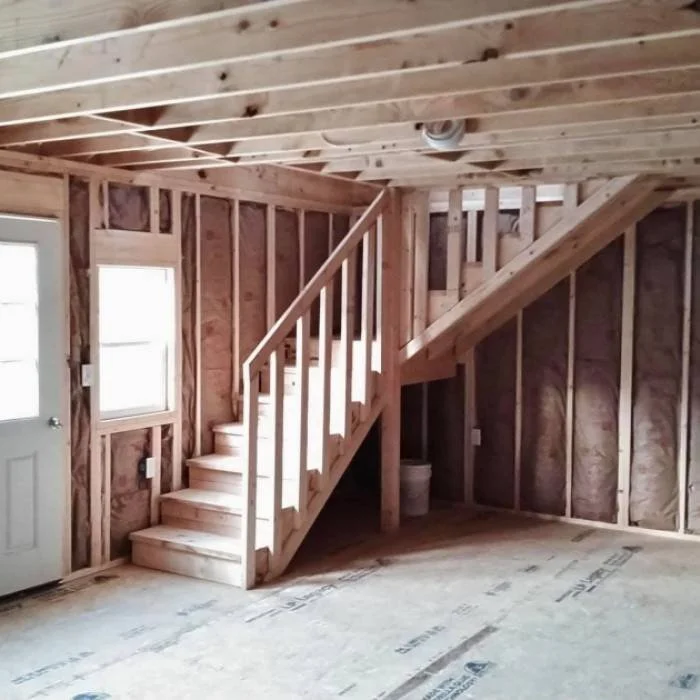 Interior view of a house under construction with exposed wooden framing, a staircase with wooden steps and railing, a window, and an unfinished floor.
