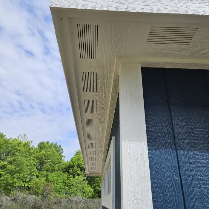 Close-up of a building's white soffit with ventilation vents, against a background of green trees and a partly cloudy sky.