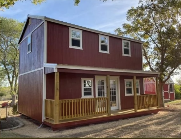 Red two-story tiny house with a wooden porch and brown railing, surrounded by trees and a clear blue sky.