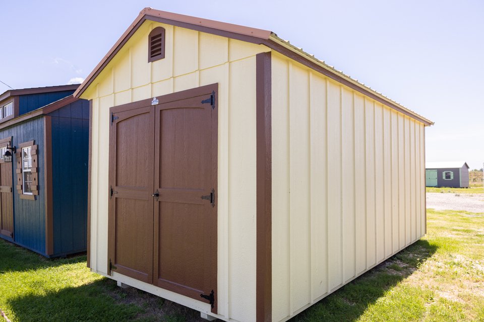 A beige metal storage shed with brown double doors, brown trim, and a small vent near the roof, situated on a grassy area under a clear sky.